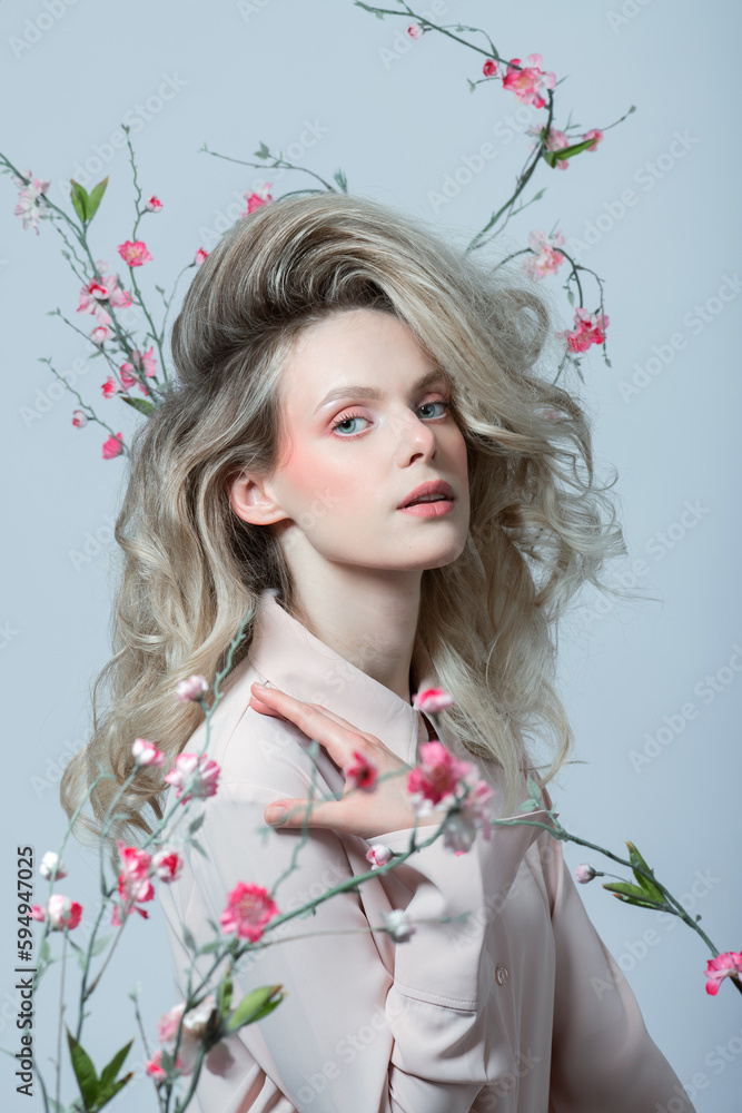 Fototapeta premium Beautiful woman with big wavy hair and purple make-up studio portrait. Model surrounded with flower twigs with blossoms and dressed with shirt looking at camera. Toned image with blue color