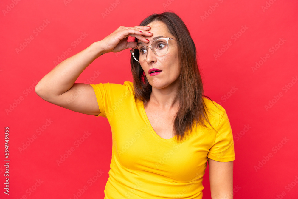 Middle-aged caucasian woman isolated on red background doing surprise gesture while looking to the side Middle-aged caucasian woman isolated on red background doing surprise gesture while looking to the side