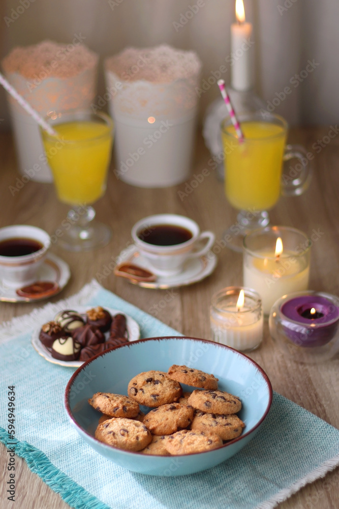 Plate of chocolate pralines, bowl of cookies, cups of tea, glasses of juice and lit candles on the table. Selective focus. Plate of chocolate pralines, bowl of cookies, cups of tea, glasses of juice and lit candles on the table. Selective focus.
