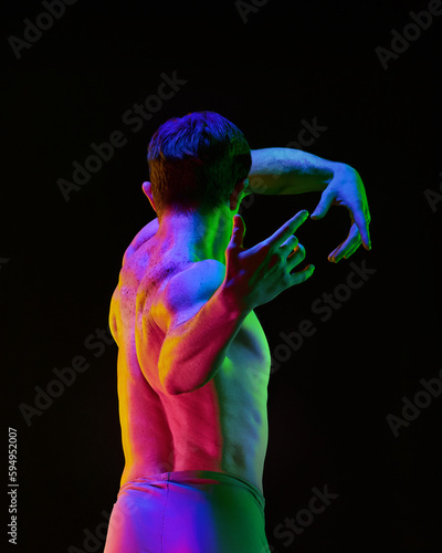 Contemporary art ballet. Young flexible athletic man, dancer posing with hands up on dark studio background with neon light. Male torso in neon glow