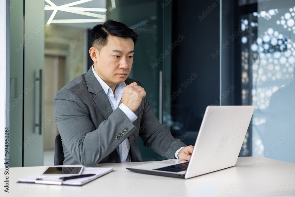 Serious thinking asian businessman working inside office sitting at table with laptop, boss making important financial decision, asian mature man in business suit at workplace.