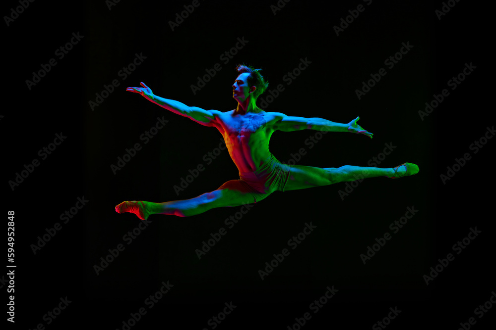 Portrait of young handsome, muscle men, ballet dancer in pastel color clothes jumping over dark background with neon light. Contemporary dance style. Split jump