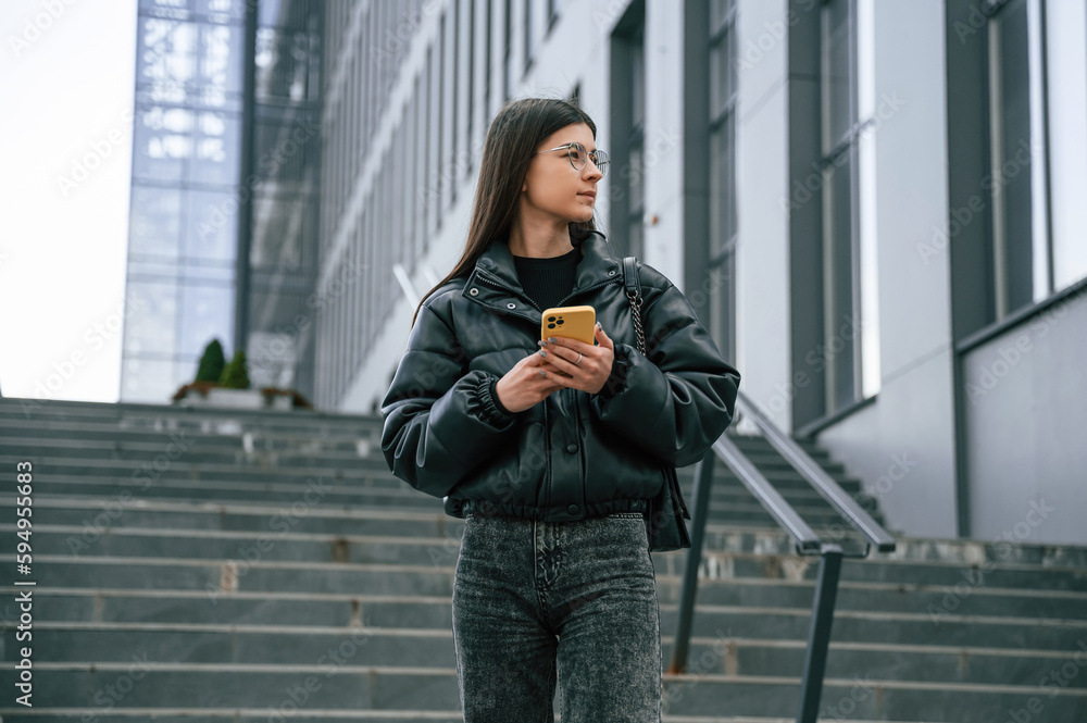 Standing on the stairs. Young woman is outdoors near building is holding smartphone Standing on the stairs. Young woman is outdoors near building is holding smartphone