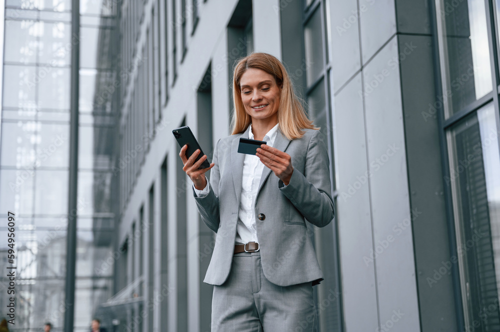 Looking at credit card, holding smartphone. Woman in formal clothes is outdoors near the business building Looking at credit card, holding smartphone. Woman in formal clothes is outdoors near the business building