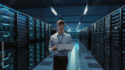 Futuristic Concept: Big Data Center Chief Technology Officer Using Laptop Standing In Warehouse, Information Digitalization Lines Streaming Through Servers. SAAS, Cloud Computing, Web Service