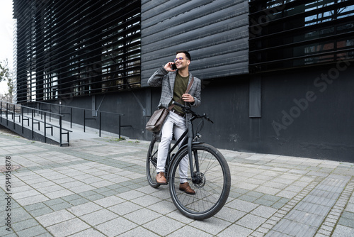 Handsome stylish businessman talking on the phone and driving a bike in the city. Businessperson in front of office building using smartphone on bicycle arrange meeting or date on lunch break