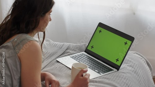 A long-haired brunette lies on a bed with a cup of tea and looks at something on her laptop. A woman lies in front of a laptop with a green screen and periodically presses the keys. photo