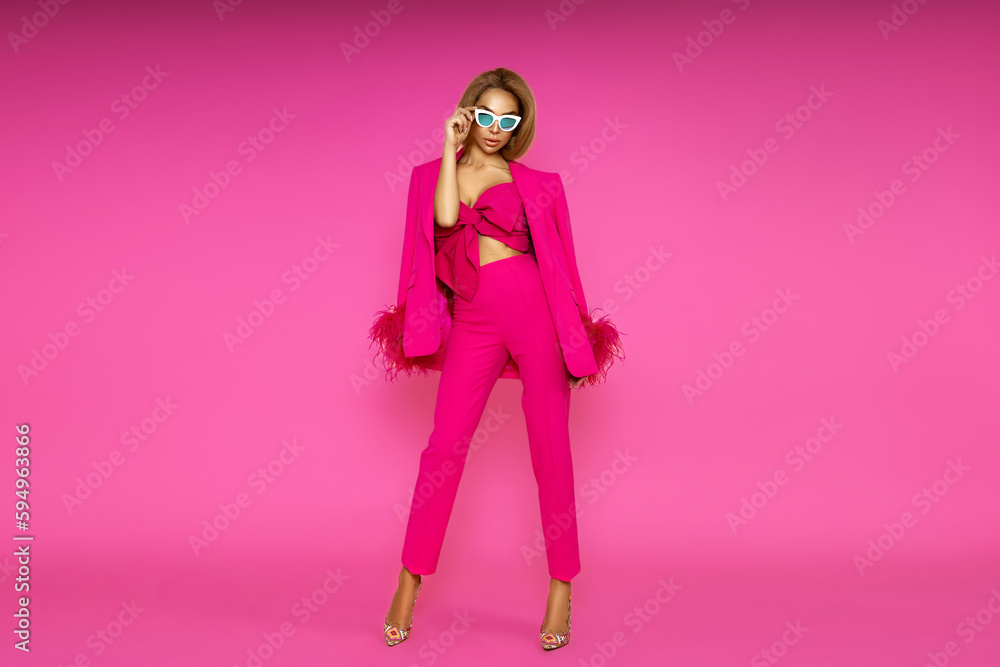 High fashion model. Elegant woman is wearing pink feather  jacket, trousers and high heels on pink background.  Chic female model posing in stylish luxury outfit.