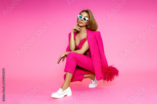 High fashion model. Elegant woman is wearing pink feather  jacket, trousers and high heels on pink background.  Chic female model posing in stylish luxury outfit.