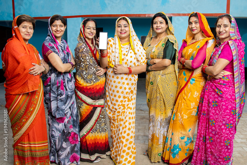Group of happy young traditional indian women housewives wearing colorful sari showing smart phone with blank display screen to put advertisement, Rural india. women empowerment.