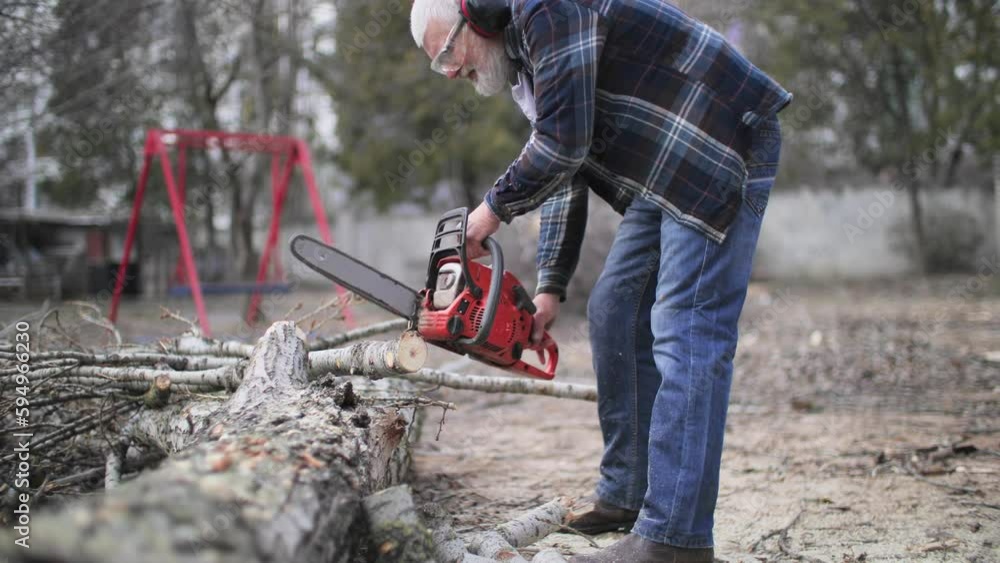 elderly gray-haired man in good health in headphones and glasses saws firewood with a chainsaw on street elderly gray-haired man in good health in headphones and glasses saws firewood with a chainsaw on street