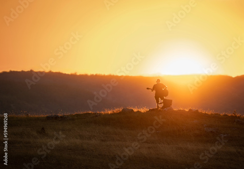 Silhouette of a man playing guitar at sunset in the field. Hills in the background. Orange colored sky.