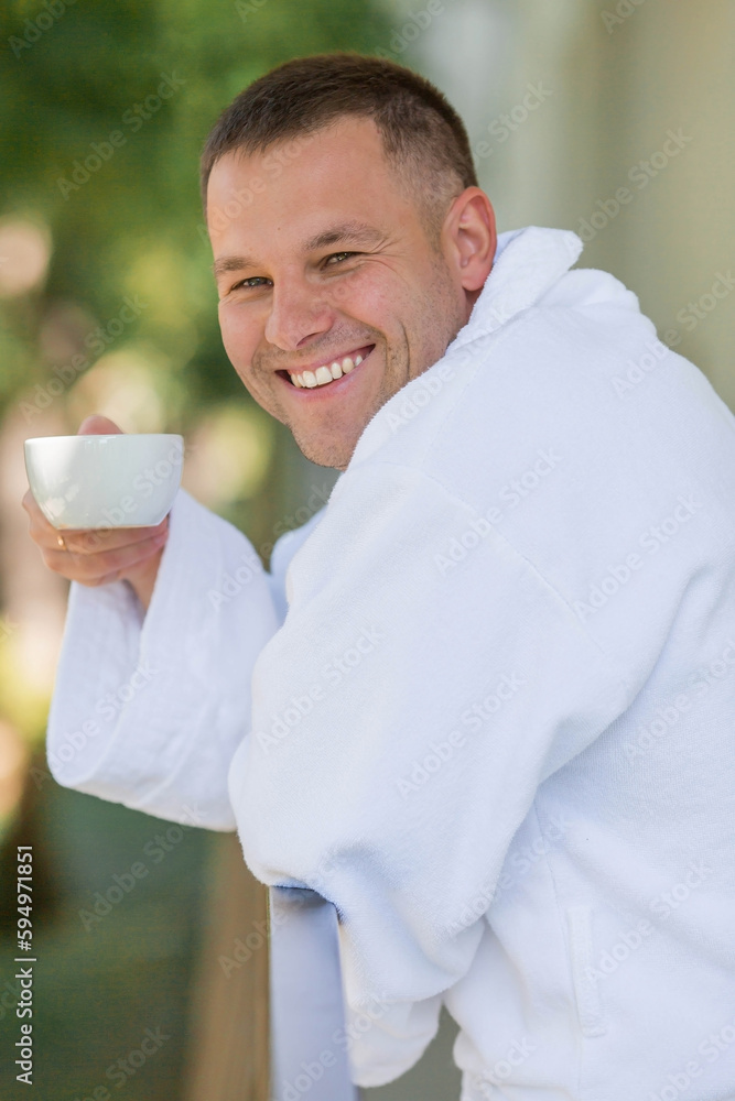 A cup of hot tea in the hands of a handsome smiling man 30+ in a white coat. Morning of the groom. The concept of hotel accommodation and the start of the wedding day. A cup of hot tea in the hands of a handsome smiling man 30+ in a white coat. Morning of the groom. The concept of hotel accommodation and the start of the wedding day.