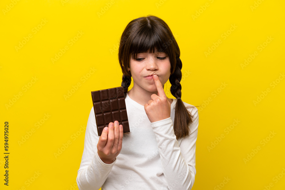 Little caucasian girl isolated on yellow background taking a chocolate tablet and having doubts Little caucasian girl isolated on yellow background taking a chocolate tablet and having doubts