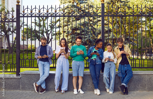 Group of young people using mobile phones on urban street. Several multiracial hipsters hanging out together, standing by beautiful city fence, holding cellphones, browsing Internet and text messaging