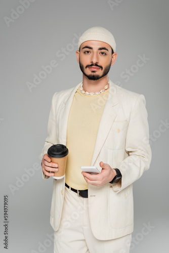 Stylish homosexual man in suit holding smartphone and coffee to go isolated on grey. © LIGHTFIELD STUDIOS