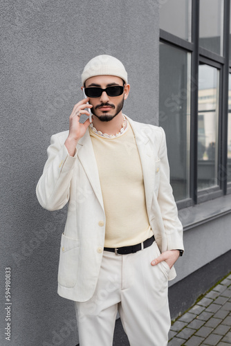 Trendy gay man in sunglasses and beige suit talking on smartphone outdoors.