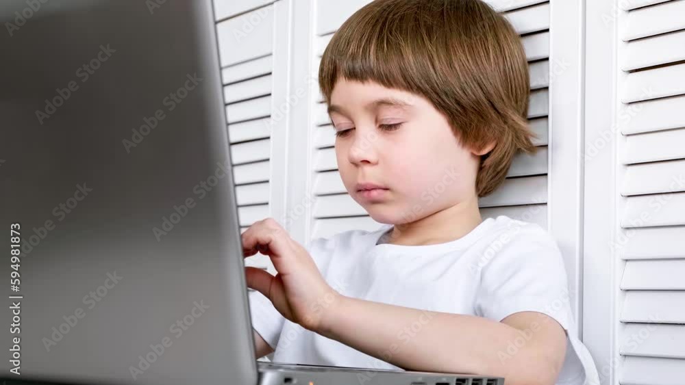 Clever 5 year boy sit at the table, uses laptop, looks at screen. Child is doing homework lesson, studying IT task, listening teacher. Home distance online e-learning concept. Wireless internet tech.
