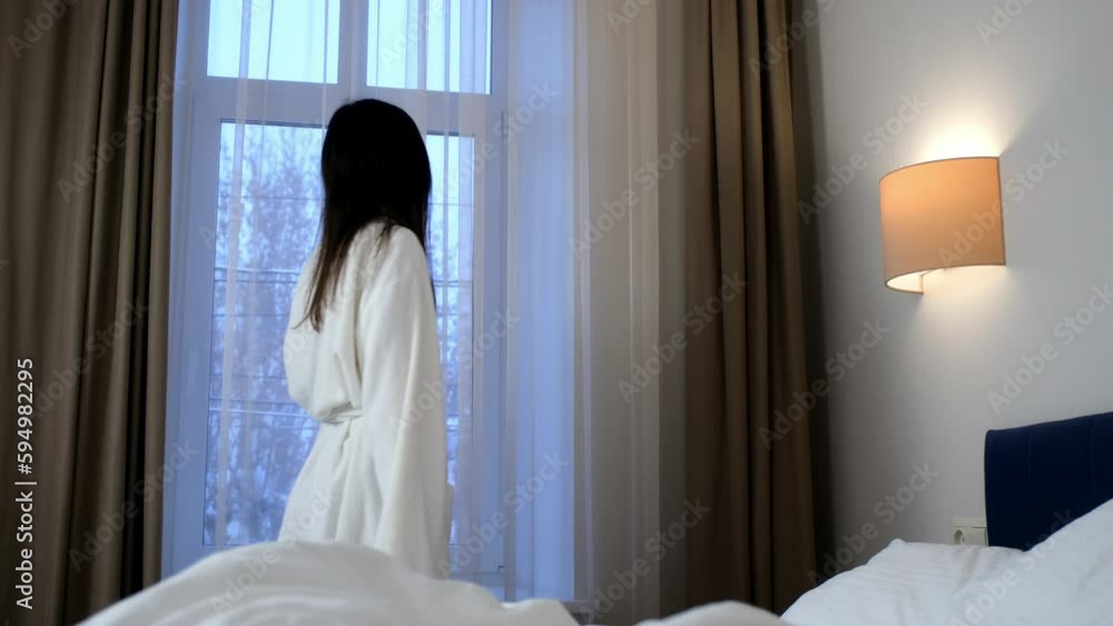 Brunette young slim woman in white bathrobe is looking at window on winter trees in hotel room. She is smiling and standing near the bed. Vaction, relax and rest with comfort in winter concept. Brunette young slim woman in white bathrobe is looking at window on winter trees in hotel room. She is smiling and standing near the bed. Vaction, relax and rest with comfort in winter concept.