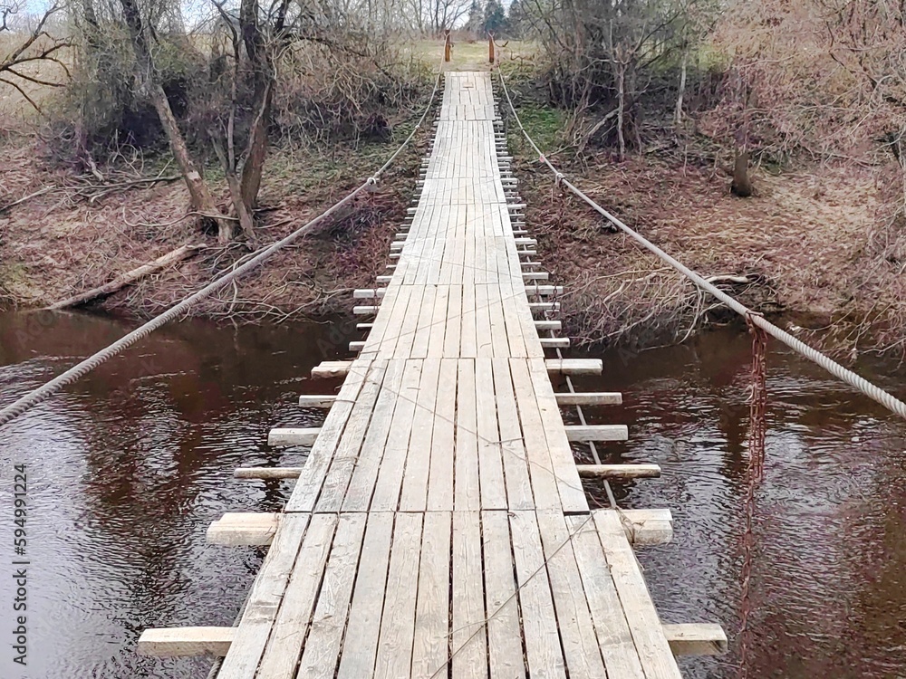 Fototapeta premium Suspended wooden pedestrian bridge to the other side of the river in the forest. View of the suspension bridge in the forest. 