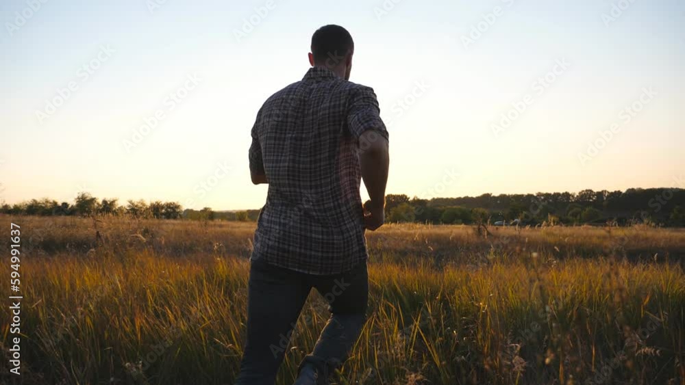 Rear view of happy man jogging through grass field with sunset at background. Young guy running among summer meadow. Male enjoying rest at beautiful nature. Concept of carefree and freedom. Close up Rear view of happy man jogging through grass field with sunset at background. Young guy running among summer meadow. Male enjoying rest at beautiful nature. Concept of carefree and freedom. Close up