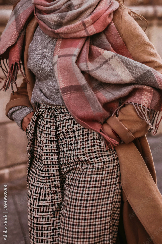 Woman in autumn stylish fashion brown long coat, scarf and plaid pants walking in the city. Female casual street style outfit