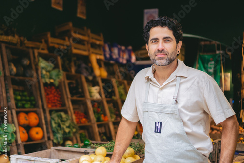 Middle-aged Latin man in an apron looking at the camera in the greengrocer's shop where he works. Copy space.