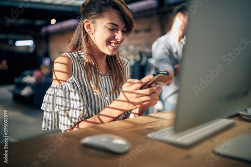 Beautiful young woman sitting at table in front of desktop computer using smartphone, feeling joyful.