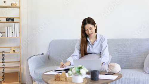 Beautiful young asian woman watching live video or video call of teacher teaching on laptop in her home  Take notes of important conversations and messages during the teacher s teaching.