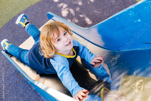 Long haired blond preschool boy plays and has fun on the slide of a playground.