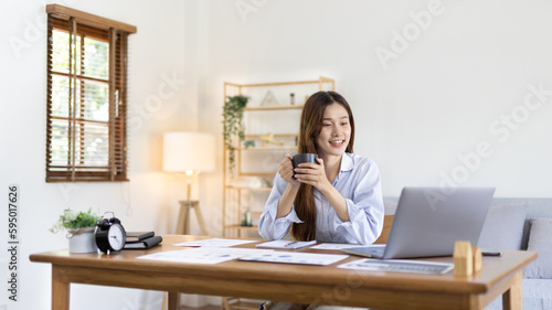 Beautiful Asian woman working on laptop and sipping coffee with smiling face in her home, Creating happiness at work with a smile, Freelancer working at home happily, Work from home. © Puwasit Inyavileart