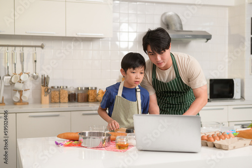 Happy young Asian father and kid are using a laptop computer to learn how to cooking food in their home kitchen.