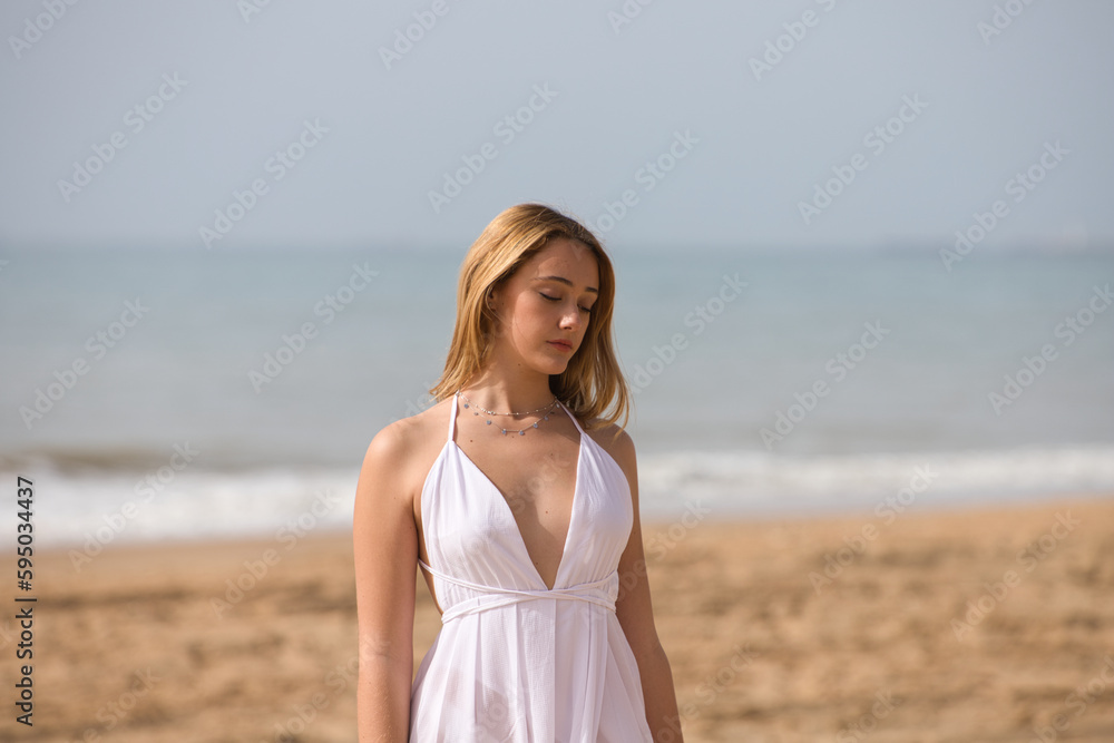 Young beautiful blonde woman in white dress is walking on the sand on the shore of the beach on a sunny day. The woman makes different body expressions. In the background the blue sea. Young beautiful blonde woman in white dress is walking on the sand on the shore of the beach on a sunny day. The woman makes different body expressions. In the background the blue sea.