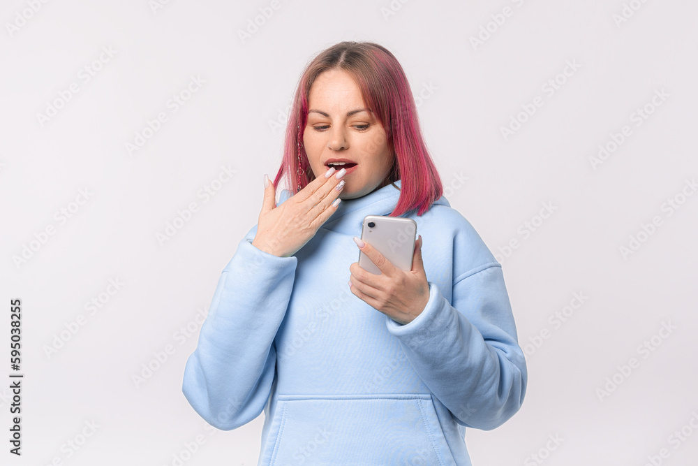 Shocked young woman looking at mobile phone, reading unexpected message with open mouth, checking social networks. Lovely female model with pink hair in a blue hoodie standing over white background Shocked young woman looking at mobile phone, reading unexpected message with open mouth, checking social networks. Lovely female model with pink hair in a blue hoodie standing over white background
