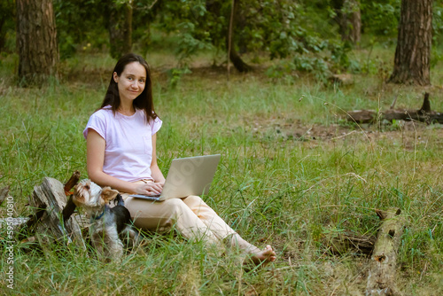 A beautiful young woman, girl with long brown hair sitting in a clearing in green forest with a little dog Yorkshire Terrier, working at laptop. Remote work, study. Student with a computer at nature.