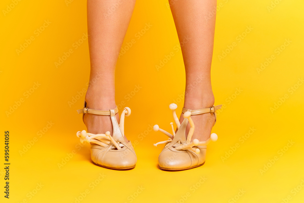 Close up portrait of beautiful female feet wearing beige heels and with mushrooms inside on yellow studio background. New spring fashion