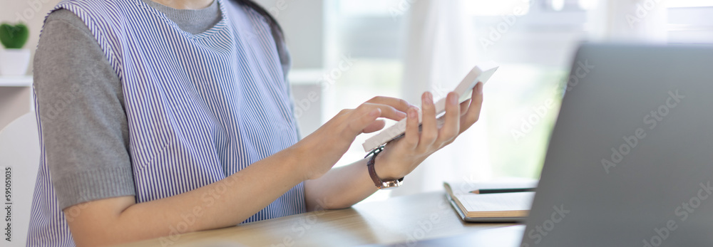 Obraz premium Asian woman working at home, Press the calculator to calculate the income - expenses of the house and use the laptop to record the information and check the accuracy, Home lifestyle.