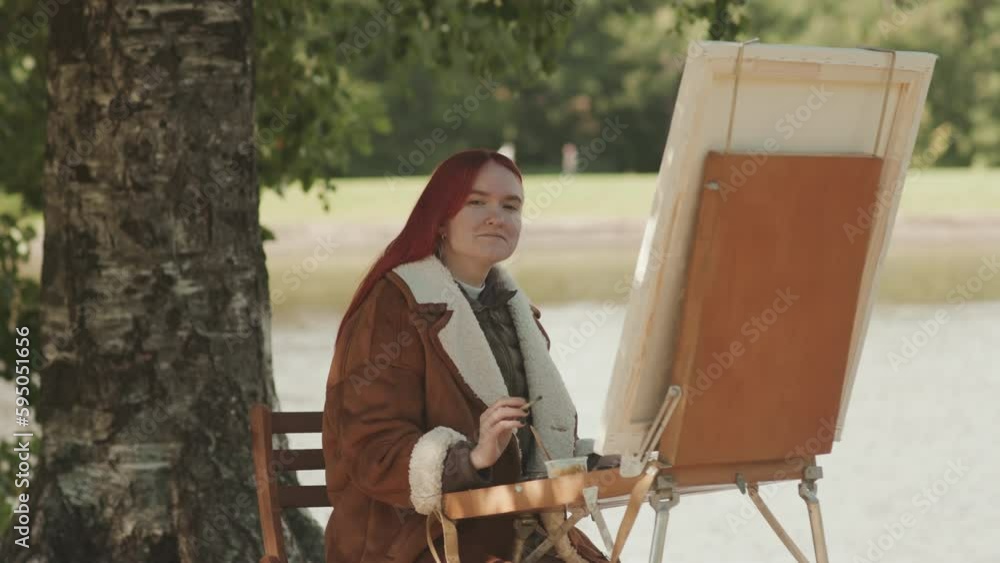 Medium portrait of Caucasian teenage girl with bright red hair smiling at camera while painting on canvas in summer park Medium portrait of Caucasian teenage girl with bright red hair smiling at camera while painting on canvas in summer park