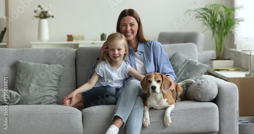 Cheerful young mother with 5s daughter and trained dog sitting on cozy couch in living room, loving parent hugging her child feeling love, enjoy happy motherhood, spend time threesome at modern home