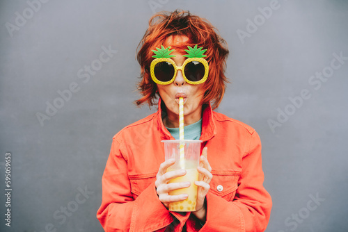 Hipster fashion young woman in bright clothes and pineapple glasses drinking fruity sugar flavored tapioca pearl bubble tea with straw on the gray wall background. Selective focus. Copy space. photo