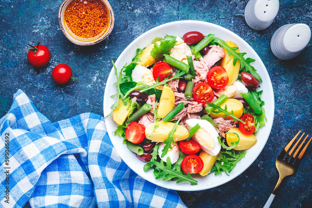 Nicoise salad with tuna, tomatoes, eggs, green beans, potatoes and olives on plate, blue table background, top view - obrazy, fototapety, plakaty 