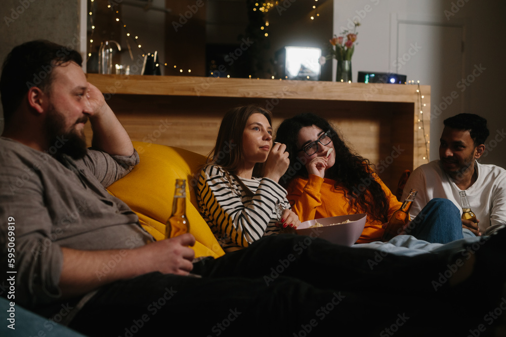 A company of multiracial friends watching a movie sitting on soft bean bags and drinking beer.