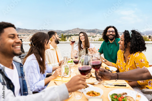 Happy multiracial young adult friends eating and drinking at barbecue party at rooftop in summer. Leisure, people and bar-b-q lifestyle