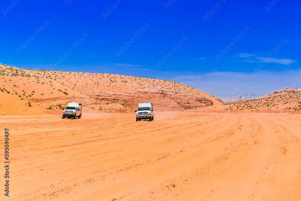 Two jeeps 4x4 moving across the sandy desert. On the way to Antelope Canyon. A picture with a blue sky and a broken unpaved road in the sand. Dusty and unpaved road through the arid and dry desert. Two jeeps 4x4 moving across the sandy desert. On the way to Antelope Canyon. A picture with a blue sky and a broken unpaved road in the sand. Dusty and unpaved road through the arid and dry desert.