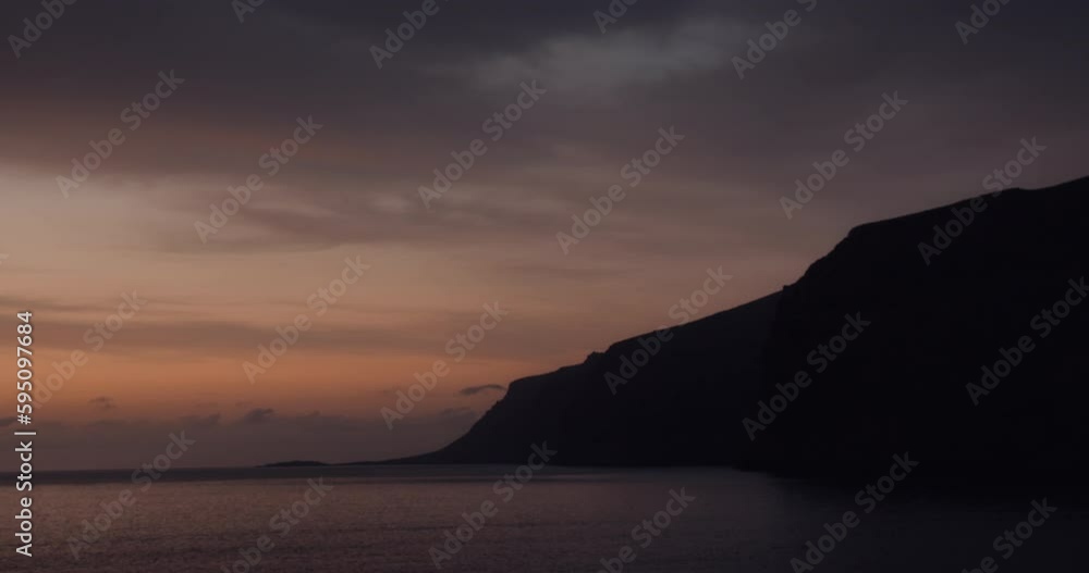 Sunset cloudy sky over dark calm ocean with mountain silhouette. Los Gigantes cliff rocks in Tenerife. Tranquil twilight seascape. Sunset cloudy sky over dark calm ocean with mountain silhouette. Los Gigantes cliff rocks in Tenerife. Tranquil twilight seascape.