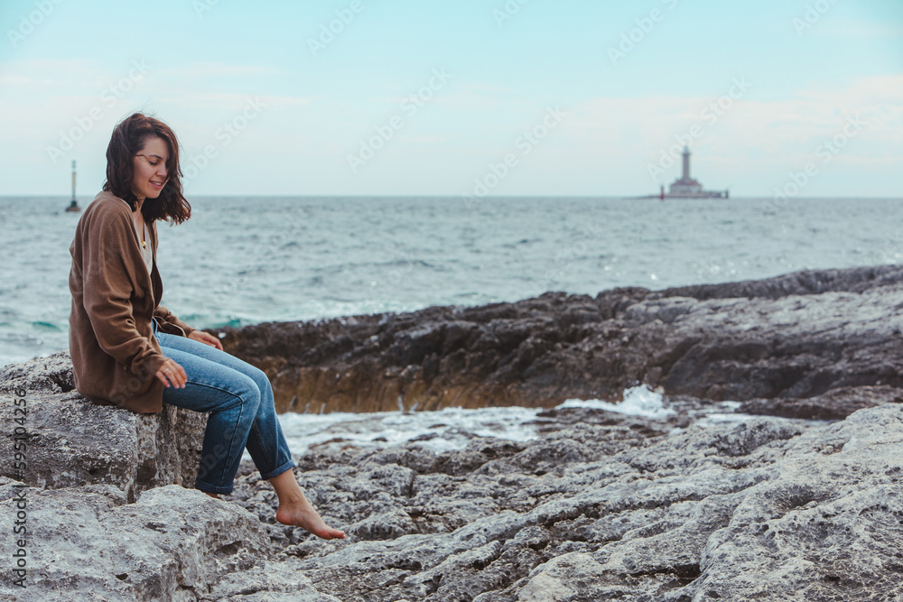 woman sitting by rocky sea beach in wet jeans lighthouse on background