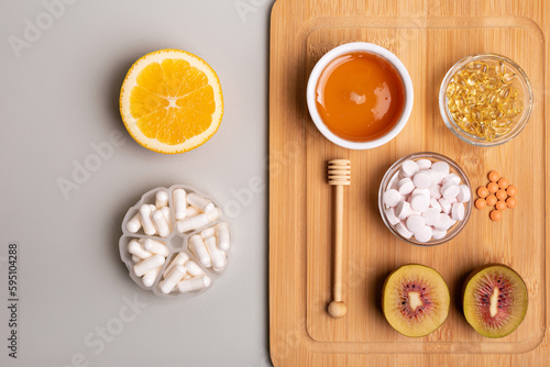 Natural vitamins from fruits and honey and vitamin pills, organic minerals in small bowls from above on wooden desk on light background. Orange, kiwi and honey as sources of natural vitamins.
