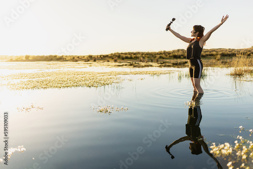 Woman taking selfie with camera in nature in middle of lake, surrounded by flowers, with selective focus, sunset in middle of lake woman traveling alone, waves in water, movie colours.