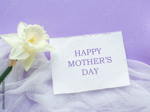 Daffodils flower on lilac background with Happy mothers day on white card. Mothers Day greeting card. Sheet of paper with text and narcissus on purple background. Congratulations note flat lay.