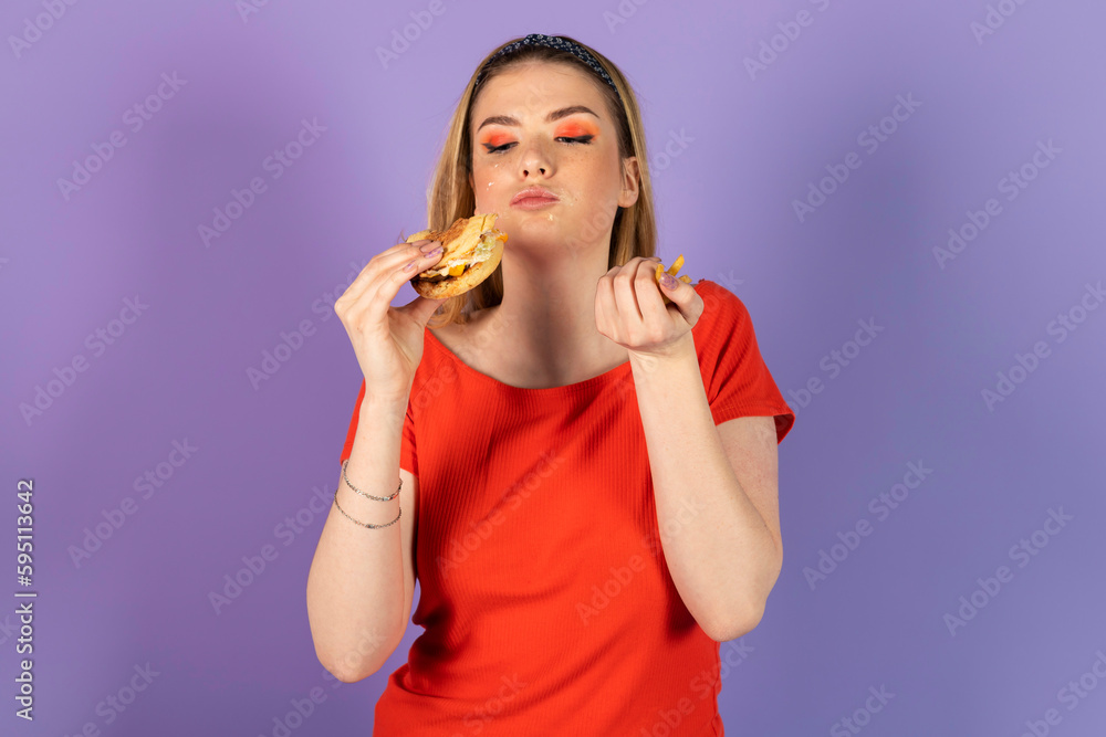 stained smeared in mayonnaise pretty girl eats fast food, a cheap burger, holds cheeseburger. fast food and unhealthy diet. isolated on purple background. stained smeared in mayonnaise pretty girl eats fast food, a cheap burger, holds cheeseburger. fast food and unhealthy diet. isolated on purple background.